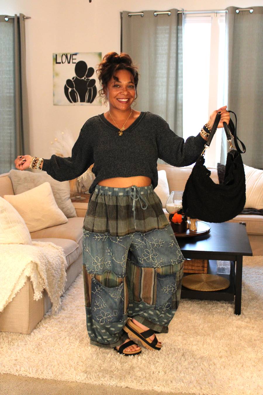 Woman in a living room wearing a dark gray sweater and blue patterned pants. Holding black fuzzy handbag.