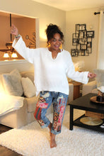 Load image into Gallery viewer, Woman in a white, soft sweater and colorful pants standing in a living room.
