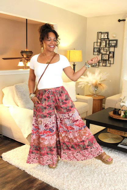 A woman modeling a pair of pink patchwork palazzo pants with abstract and floral motifs, paired with a white top, standing in a room with a white carpet and furniture.