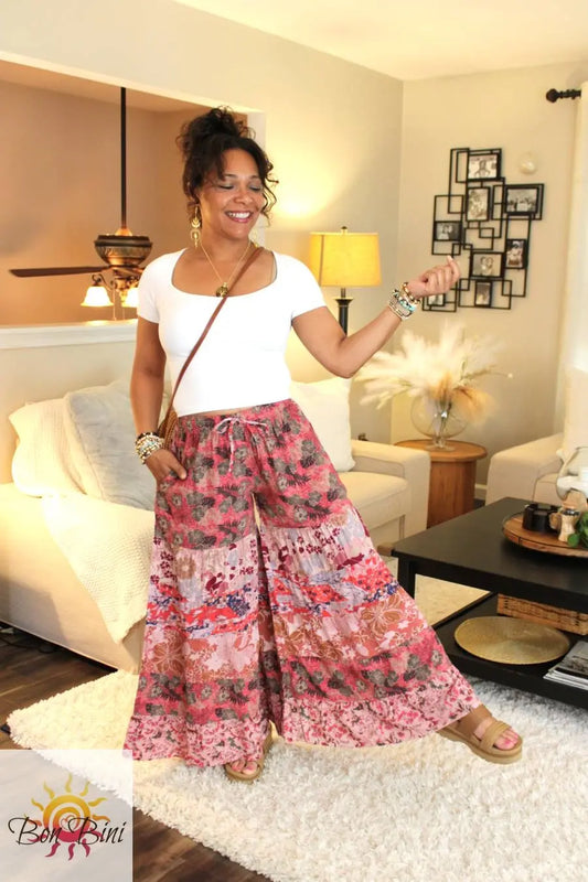 A woman modeling a pair of pink patchwork palazzo pants with abstract and floral motifs, paired with a white top, standing in a room with a white carpet and furniture.