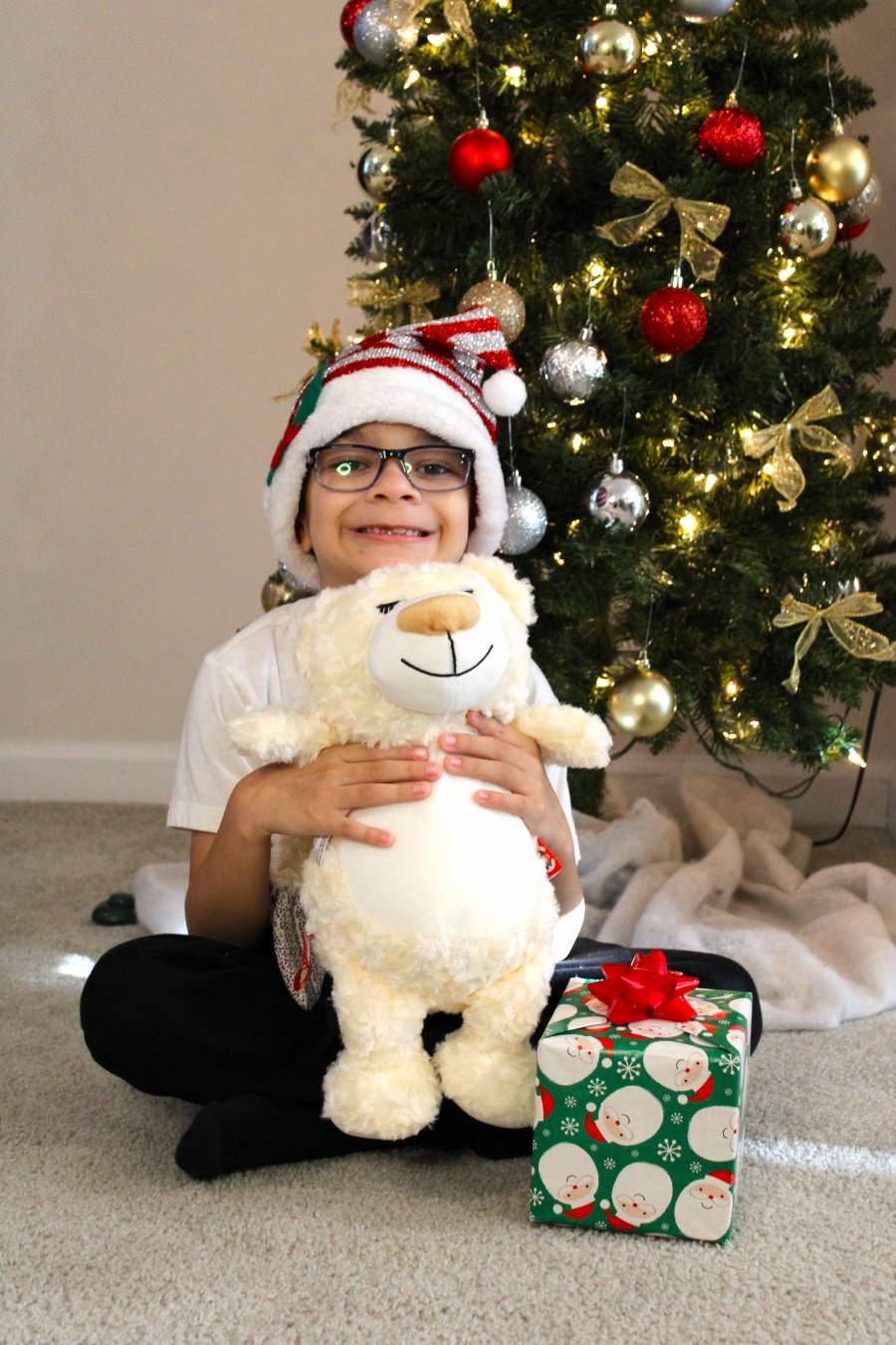 Child holding a teddy bear in front of a Christmas tree with ornaments and a gift.